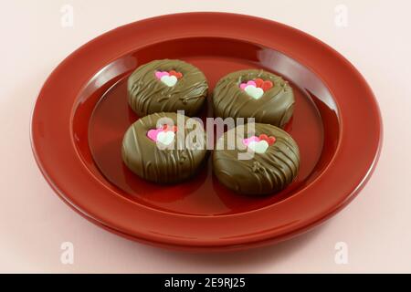 Valentine`s Day chocolate dipped sandwich cookies with heart decorations on red plate on pink background Stock Photo